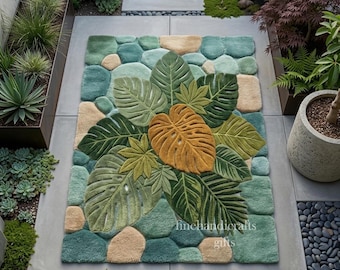 Puede incluir: Una alfombra rectangular con un diseño de hojas tropicales. La alfombra presenta hojas grandes y detalladas en tonos verdes y una hoja central de color amarillo dorado. El fondo tiene un patrón de formas turquesas y beige, que se asemejan a piedras.