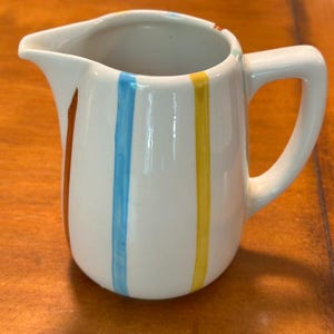 May include: A white ceramic creamer pitcher featuring a series of vertical hand-painted stripes in shades of orange, light blue, and yellow. The pitcher has a curved pouring spout and a sturdy side handle, with a smooth, glossy finish throughout.