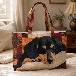 May include: A tote bag featuring a patchwork design in shades of red, orange, and brown. The front displays a detailed illustration of a black and tan dachshund sleeping on a beige pillow. The bag has two matching fabric handles at the top.