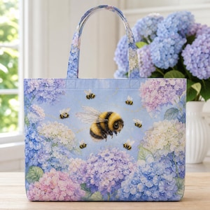 Op de afbeelding: Een rechthoekige stoffen draagtas met een lichtblauwe achtergrond en een printpatroon van paarse, blauwe en witte hortensiabloemen. Een grote, gedetailleerde hommel staat centraal op de tas, omringd door verschillende kleinere hommels.