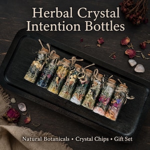May include: A set of eight clear glass bottles filled with natural botanicals and crystal chips, each sealed with a cork and twine. The bottles feature small metal charms and are arranged on a black tray. Text reads: Herbal Crystal Intention Bottles.