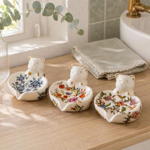 Könnte beinhalten: Drei herzförmige Schmuckschalen aus Keramik, jede mit einer kleinen weißen Bärenfigur an der Rückseite. Die erste Schale hat ein blaues Blumenmuster, während die zweite und dritte Schale bunte botanische Drucke mit Blumen und Früchten zeigen.