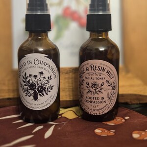 以下が含まれることがあります： 黒いスプレーキャップが付いた2つの茶色のガラススプレーボトル。ボトルには、黒い文字と花の模様が入った白いラベルが付いています。1つのラベルには「Rooted in Compassion」、もう1つには「Rose & Resin Mist Facial Toner」と書かれています。