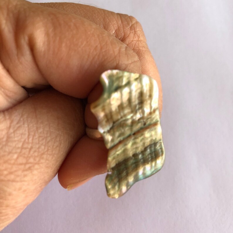 May include: A ring made from a large, iridescent seashell with a natural, irregular shape. The shell is a mix of green, brown, and gold tones.
