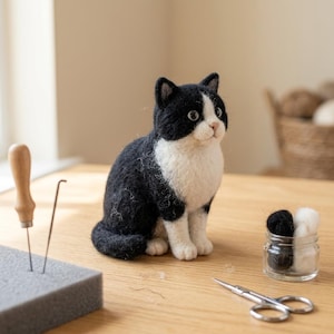 May include: A needle-felted black and white cat figurine sitting upright. The cat has a black back, head, and tail with a white chest, face, and paws. Nearby are a felting needle, a gray foam block, a small glass jar with wool, and scissors.