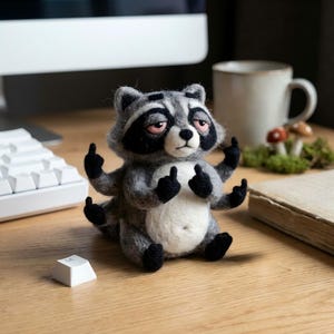 Puede incluir: Figura de mapache de fieltro hecha a mano con pelaje gris y blanco, patas negras y una máscara negra alrededor de los ojos. El mapache está haciendo la señal del dedo medio con sus seis manos. La figura está sobre un escritorio de madera cerca de un teclado y una taza de café.