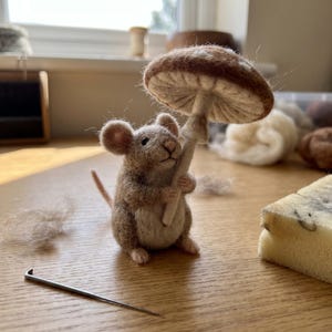 May include: A needle-felted mouse figurine made of brown and tan wool, holding a large, textured brown and cream mushroom. The mouse has small round ears, a thin tail, and a light-colored belly. A single metal felting needle lies nearby.