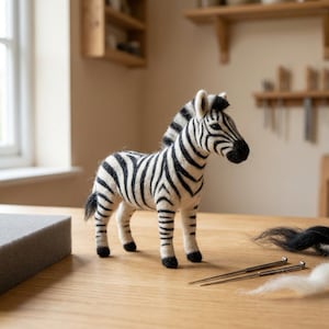May include: A needle-felted zebra figurine featuring a white body with distinct black stripes, a black mane, and a black tail. The zebra stands on four legs with black hooves, facing toward the right side of the frame, crafted with a textured wool finish.