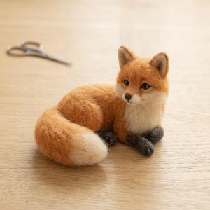 May include: A needle-felted fox figurine with orange and white fur, dark gray paws, and a bushy tail with a white tip. The fox is in a resting position with its head turned to the side. A pair of metal scissors is visible in the background.