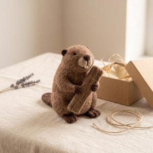 May include: A needle-felted brown beaver figurine holding a small wooden log. The beaver has a textured, fuzzy appearance with dark eyes and visible front teeth. It is positioned next to an open cardboard gift box and a piece of twine.