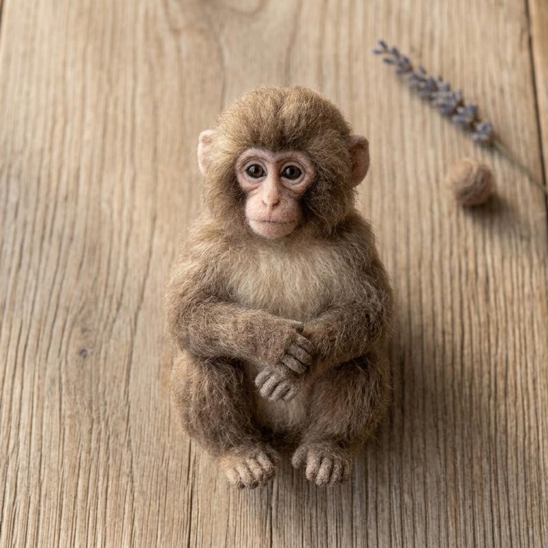 May include: A needle-felted monkey figurine with textured brown and tan fur, dark eyes, and small ears. The monkey is in a seated position with its hands resting on its lap, featuring detailed facial features and a soft, fuzzy appearance.