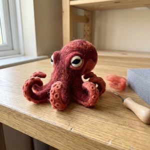 May include: A needle-felted octopus figurine in a deep reddish-brown hue, featuring a textured surface, prominent white and black eyes, and curled tentacles with visible suction cup details, accompanied by a small tuft of loose wool and a felting needle.