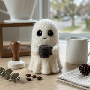 Puede incluir: Una figura de fantasma de fieltro blanco hecha a mano sosteniendo una taza de café marrón oscuro. El fantasma tiene grandes ojos negros y una pequeña sonrisa. También se ven granos de café y una jarra blanca.