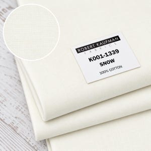 May include: Folded white 100% cotton fabric by Robert Kaufman Fabrics, labeled K001-1339 Snow. The material features a plain weave texture, shown in a circular inset detail, with a clean, solid white color throughout the textile layers.
