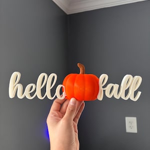 May include: A three-dimensional sign featuring the white cursive text hello and fall with a bright orange pumpkin replacing the letter o. The pumpkin has a brown stem and a rounded, ribbed texture. The entire piece is constructed as one unit.