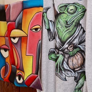 Può includere: Un cuscino quadrato con un design astratto e colorato di un volto con forme geometriche in rosso, giallo, blu e viola, accanto a una t-shirt grigio chiaro che mostra un camaleonte verde che indossa una veste e tiene un piccolo sacchetto.