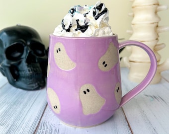 May include: A lavender ceramic mug with a handle, decorated with beige ghost shapes. The mug is filled with whipped cream, drizzled with chocolate sauce and sprinkles. A black skull and a spine decoration are in the background.