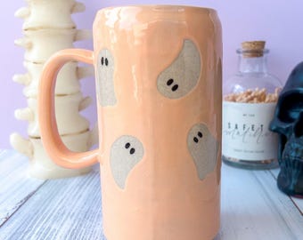 May include: A peach-colored ceramic mug with a large handle, decorated with four gray ghost designs. The mug has a glossy finish and a cylindrical shape. The background includes a decorative spine, a glass bottle, and a black skull.
