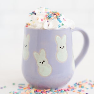 Könnte beinhalten: Eine lavendelfarbene Keramiktasse mit hellgrünen Hasenmustern. Die Tasse ist mit Schlagsahne und bunten Streuseln gefüllt. Die Tasse hat einen Henkel und steht auf einer weißen Oberfläche.