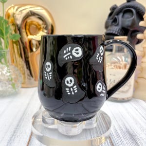 May include: Black ceramic mug decorated with white ghost illustrations. The mug has a glossy finish and a curved handle. The mug is sitting on a clear, decorative coaster. Halloween-themed drinkware.