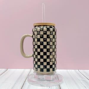 Puede incluir: Una taza de cerámica con un patrón de cuadros blancos y negros, un asa beige y una tapa de bambú. Una pajita de cristal transparente está insertada en la tapa. La taza se apoya en un posavasos decorativo transparente.