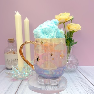 Puede incluir: Una taza de cerámica iridiscente con asa dorada, decorada con nubes y estrellas, llena de algodón de azúcar azul. Descansa sobre un posavasos transparente con forma de flor. Flores amarillas y velas en el fondo.