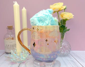 May include: A ceramic mug with a gold handle and iridescent finish, featuring a cloud and star design. The mug is filled with blue cotton candy and sits on a clear, flower-shaped coaster. Yellow flowers and candles are in the background.