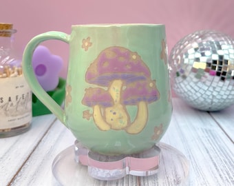 May include: A light green ceramic mug with a large mushroom design in purple, yellow, and white. The mug has a curved handle and small flower accents. The mug is sitting on a clear, pink-accented stand. A disco ball is in the background.