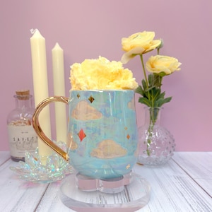 Pode incluir: Uma caneca de cerâmica azul iridescente com uma alça dourada, desenhos de nuvens e diamantes e um arranjo floral amarelo no interior. A caneca está sobre um porta-copos transparente em forma de flor. Duas velas brancas e uma garrafa de vidro pequena estão no fundo.