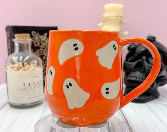 Può includere: Tazza in ceramica arancione con illustrazioni di fantasmi bianchi. La tazza ha un manico grande ed è appoggiata su un supporto trasparente e decorativo. Lo sfondo include una bottiglia di vetro, un teschio e una candela.