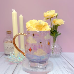 May include: A white ceramic mug with a black handle and the text "But First, Coffee" in a bold, black font. The mug is set against a blurred background. The mug appears to be approximately 10 cm tall.