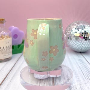 以下が含まれることがあります： 茶色とピンクの花柄が施された、取っ手付きの淡い緑色のセラミックマグ。 マグカップは、透明でピンクの花の形をしたコースターの上に置かれています。 ディスコボールやその他の装飾品が背景にあります。