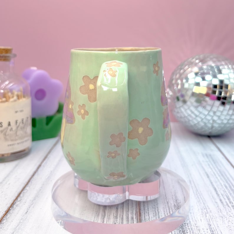 以下が含まれることがあります： 茶色とピンクの花柄が施された、取っ手付きの淡い緑色のセラミックマグ。 マグカップは、透明でピンクの花の形をしたコースターの上に置かれています。 ディスコボールやその他の装飾品が背景にあります。