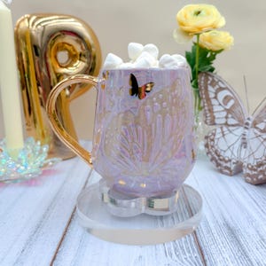 May include: A pearlescent light purple mug with a gold handle, featuring embossed butterfly wing patterns and a small black butterfly decal. The mug is filled with white marshmallows and sits on a clear, flower-shaped acrylic coaster.
