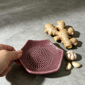 Könnte beinhalten: Eine mauvefarbene Keramikreibe mit strukturierter Oberfläche. Ingwer, eine ganze Knoblauchknolle und zwei Zehen liegen neben der Reibe. Die Reibe ist etwa 15 cm lang.