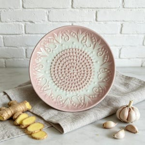 May include: A pink and white ceramic grater plate with a textured center and decorative floral border. Ginger slices, a whole garlic bulb, and garlic cloves are arranged around the plate on a linen cloth.
