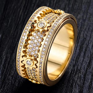 May include: A gold-colored ring with a unique gear design. The ring features multiple rows of small, sparkling clear stones and three gear-shaped accents with a single clear stone in the center of each. The ring is set against a dark background.