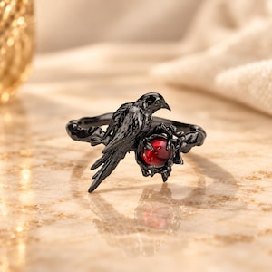 May include: A black metal ring featuring a sculpted bird perched on a branch-like band. The bird is positioned next to a small, round, translucent red gemstone held within a dark, textured, organic-shaped setting.