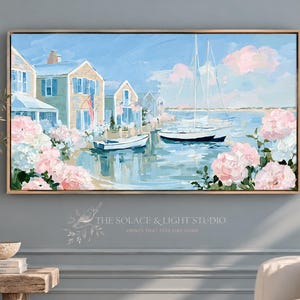 Könnte beinhalten: Ein rechteckiges gerahmtes Gemälde zeigt eine Küstenszene mit hellen Häusern, Segelbooten auf ruhigem blauem Wasser und großen rosa Blumenbüscheln im Vordergrund. Der Text THE SOLACE & LIGHT STUDIO PRINTS THAT FEEL LIKE HOME ist zu sehen.