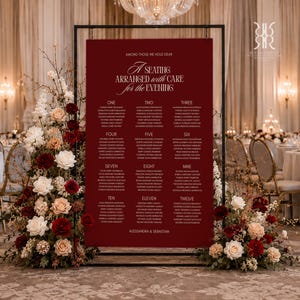 May include: A burgundy seating chart with elegant calligraphy, framed in black, for a wedding reception. The chart lists guest names under numbers one through twelve. Floral arrangements of white, red, and peach roses flank the chart, creating a romantic ambiance.