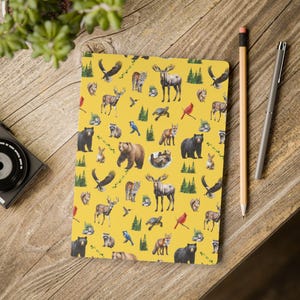 Peut inclure: Un carnet rectangulaire à couverture jaune orné d'un motif répétitif d'animaux sauvages nord-américains, notamment des orignaux, ours noirs, cerfs, renards, écureuils, tortues et oiseaux, parsemé de petites illustrations de pins verts.