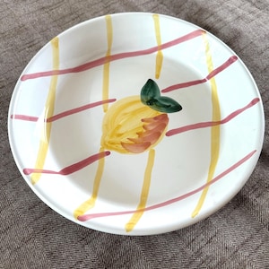 May include: White ceramic plate with a hand-painted lemon design. The plate features a yellow and pink plaid pattern. The lemon has yellow and brown tones with a green leaf. A decorative piece for the kitchen or dining room.