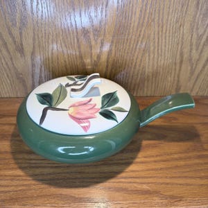 May include: A green ceramic serving dish with a lid. The lid is white with a floral design in pink, green, and brown. The dish has a handle and is set on a wooden surface. The dish is likely vintage.