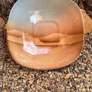 May include: A square, shallow bowl with a gradient color scheme. The bowl transitions from a light blue-gray at the top to a warm orange-brown at the bottom. The design resembles a landscape with layered, horizontal bands.