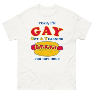 Peut inclure: T-shirt blanc avec un dessin de hot-dog et le texte "Yeah, I'm GAY Got A Yearning For Hot Dogs" en rouge et bleu. Le hot-dog est jaune avec des accents rouges et jaunes.