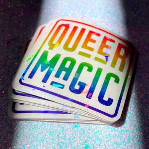 May include: A stack of rainbow holographic stickers with the text "Queer Magic" in a white square.