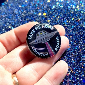 May include: A black and white button with a silver UFO and the text "Take Me Home Country Roads".