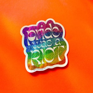 May include: A rainbow glitter sticker with the text "pride was a riot" in white letters. The sticker is on a bright orange background.