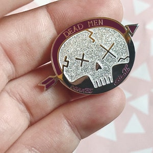 May include: A silver and burgundy enamel pin with a skull design. The skull is white with a glittery finish and has two crossed bones for eyes. The text "DEAD MEN CANNOT" is written around the skull in a burgundy enamel.