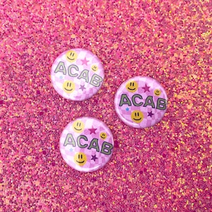 May include: Three pink and white button pins with the acronym "ACAB" and smiley faces. The pins are on a pink glitter background.